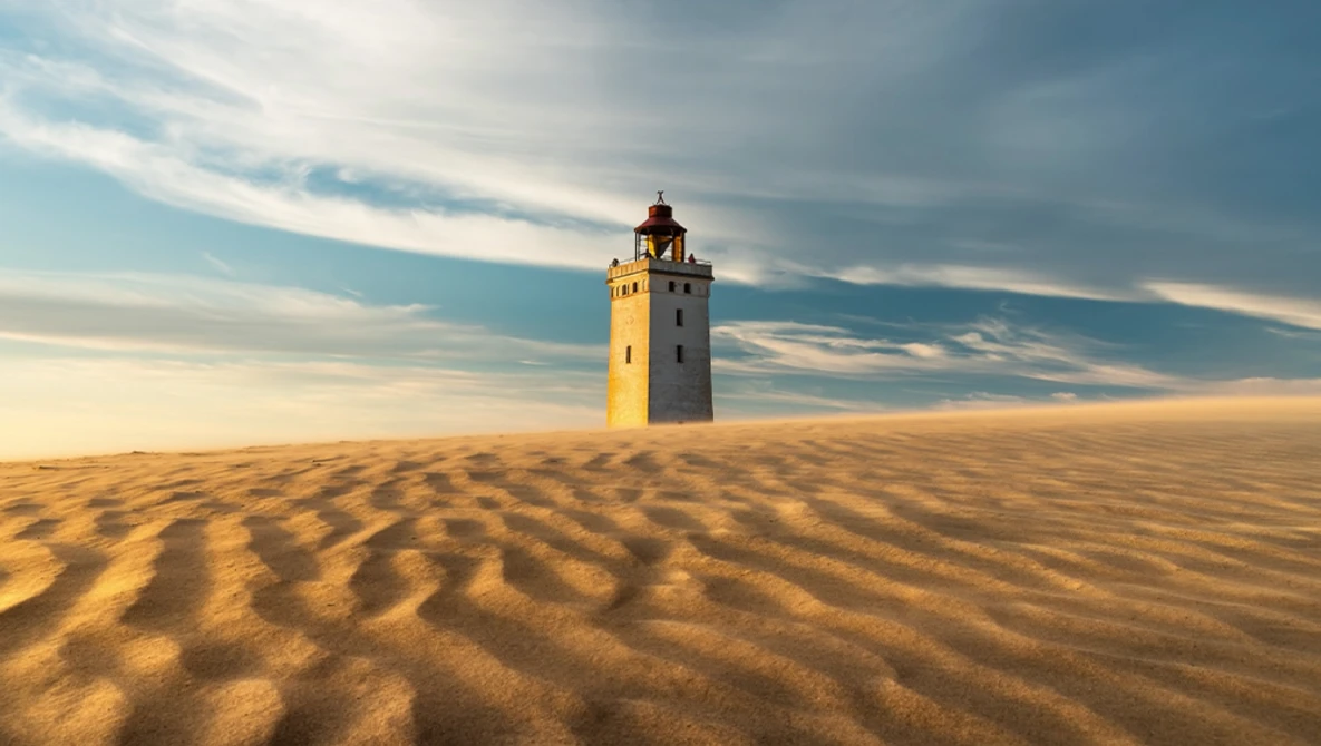 Abandoned yellow lighthouse tower standing alone in vast desert sand dunes under dramatic sky.