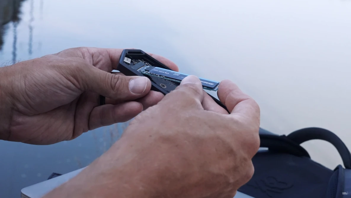 Hands holding and examining a compact camera lens or optical device against a cloudy sky backdrop.