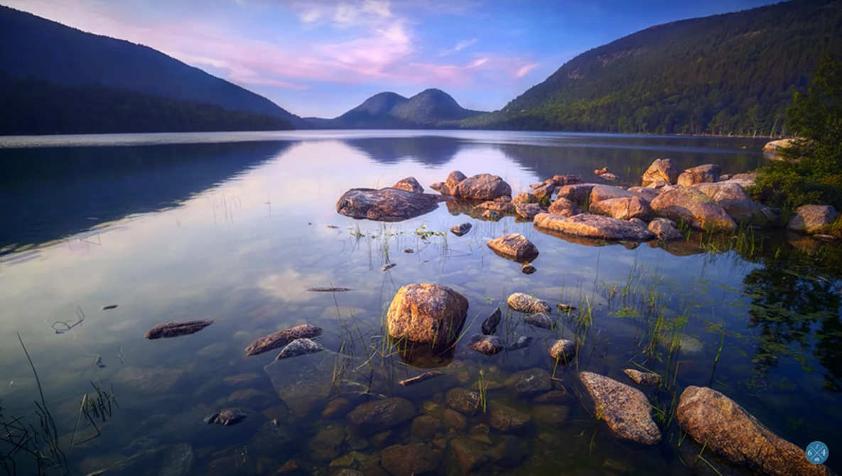 Calm alpine lake at sunrise with scattered boulders in shallow water, forested mountains in background.