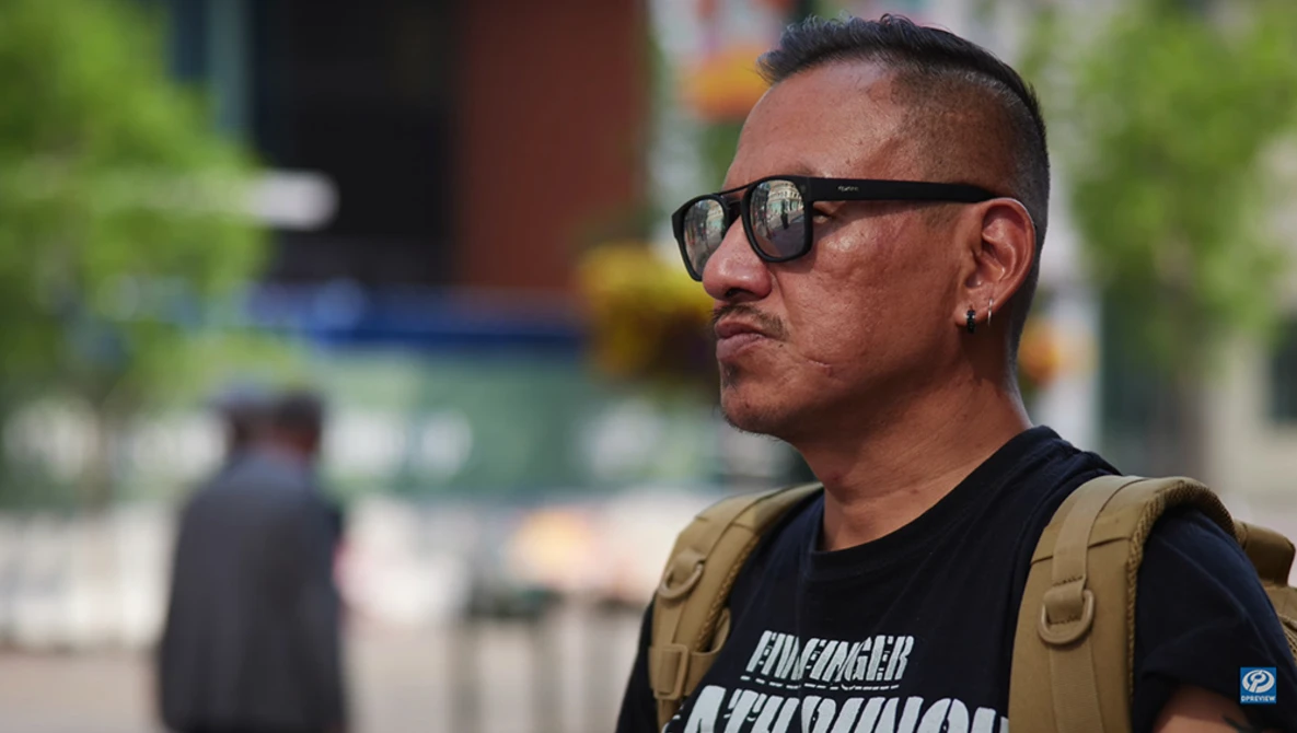 Man wearing sunglasses and tan vest standing outdoors with blurred urban background.
