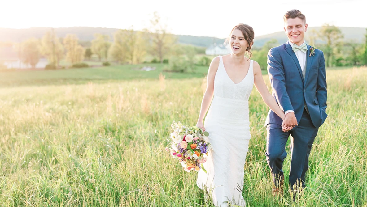 Bride and groom walking hand-in-hand through a grassy field at golden hour.