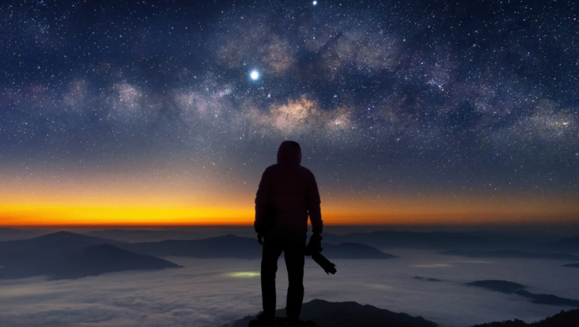 Silhouetted figure standing on a mountain peak beneath the Milky Way galaxy at twilight.