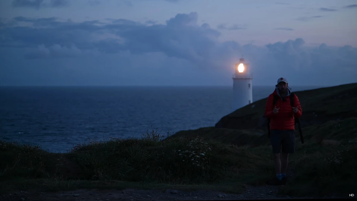 Solo hiker in red jacket standing on coastal cliff at dusk, facing illuminated lighthouse beacon on distant headland.