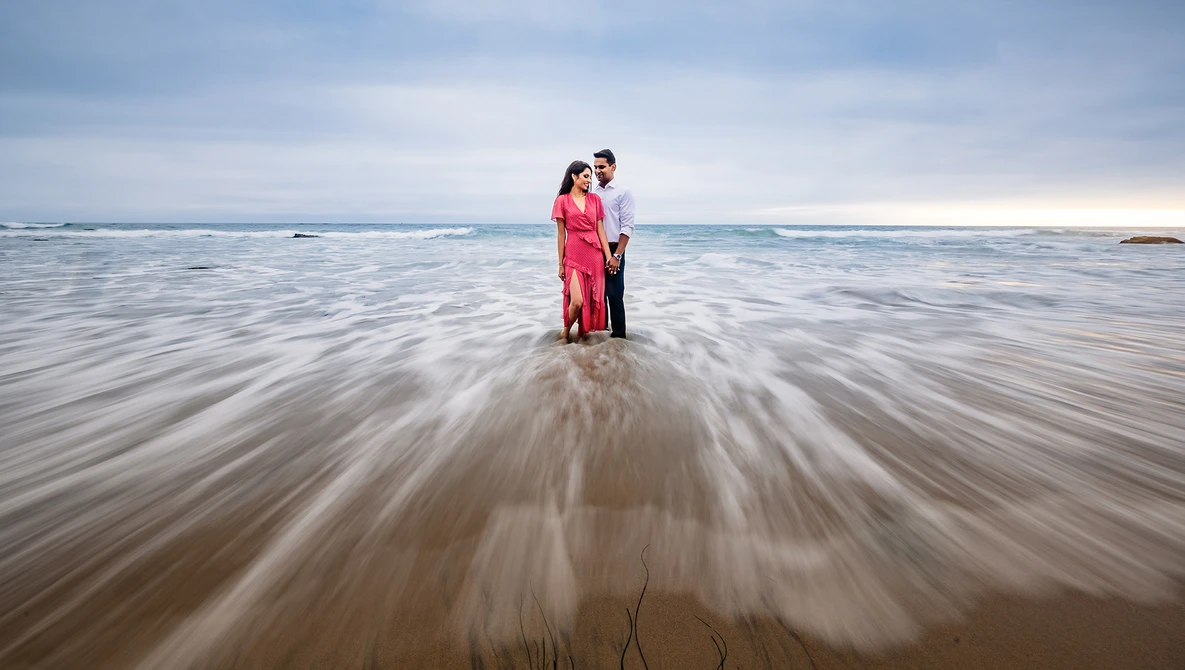 Couple standing in shallow ocean water with long exposure effect showing motion blur of waves.