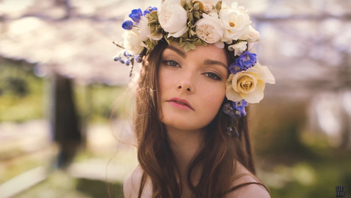 Young woman wearing an elaborate floral crown with white, cream, and blue flowers, gazing directly at camera.