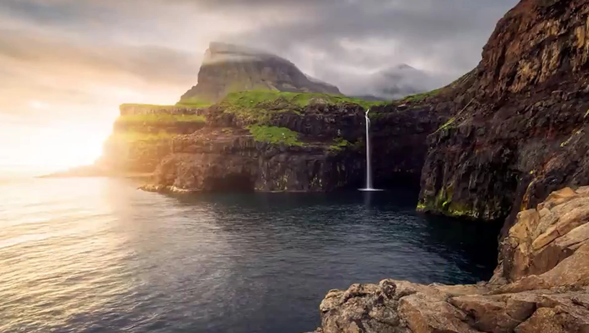 Dramatic coastal landscape with waterfall cascading into rocky cove beneath towering cliff faces at golden hour.