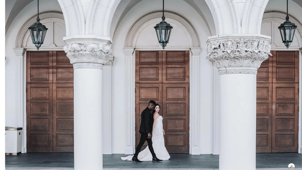 Bride and groom walking between white columns under classical arches with wooden doors.