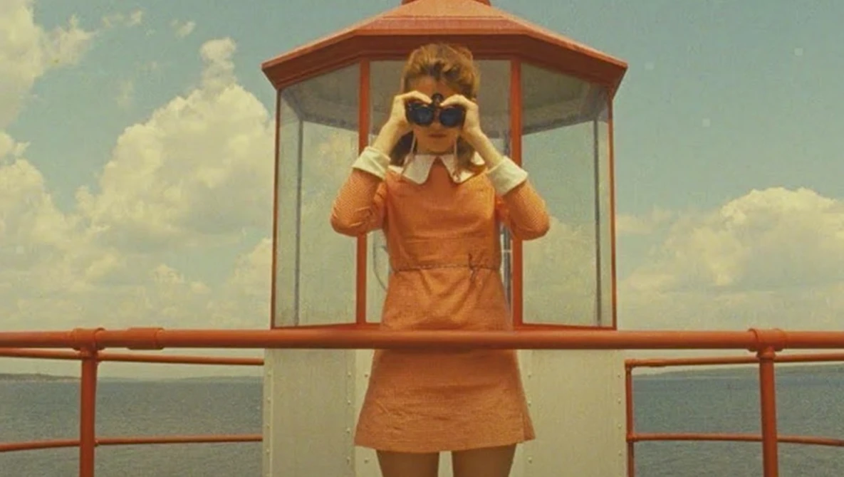 Woman in orange dress and sunglasses standing in vintage lifeguard tower overlooking calm water.