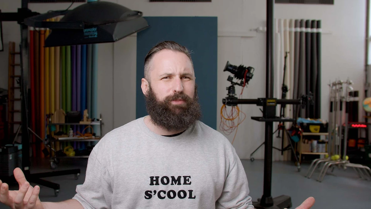 Man with beard wearing HOME S'COOL shirt in a photography studio with lighting equipment visible.