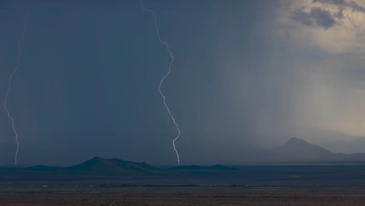 Lightning bolt streaks across stormy sky above distant mountain ranges in arid landscape.