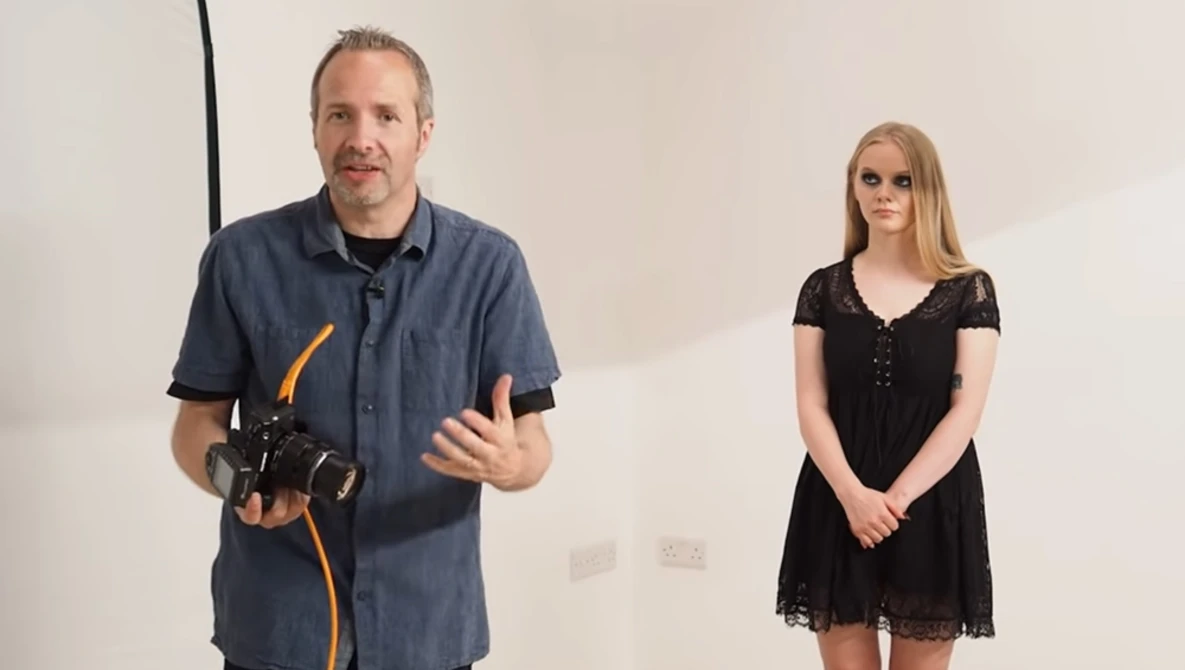 Man in navy shirt holding camera equipment gestures while speaking next to woman in black lace dress.