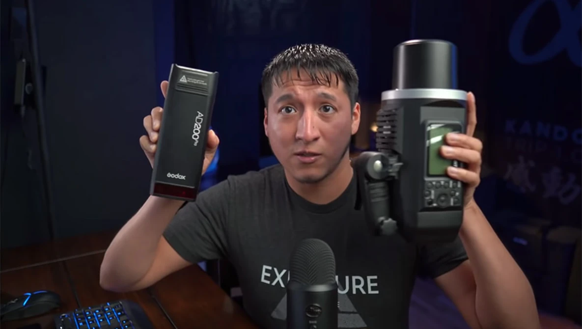 Man holding two off-camera flash units while speaking to camera in studio setting.