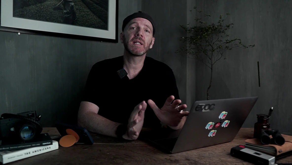 Man speaking while gesturing at laptop displaying stickers in dimly lit studio workspace.
