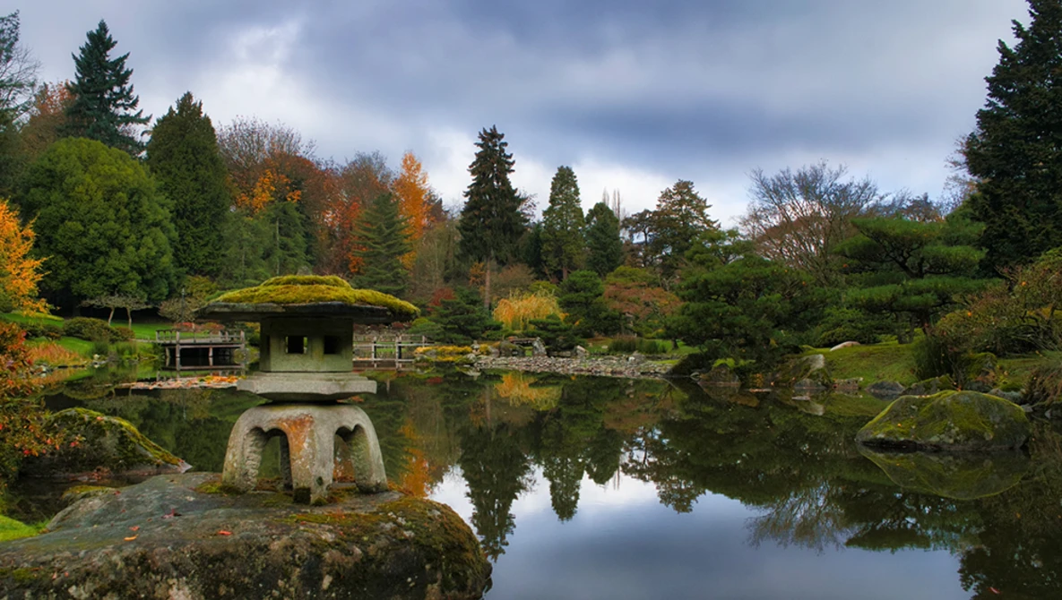 Stone lantern on moss-covered rock beside calm pond reflecting autumn foliage and evergreen trees.