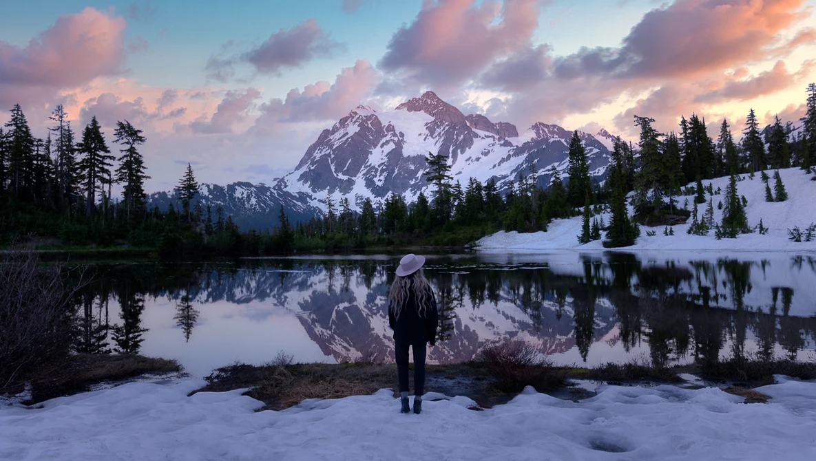 Hiker standing on snowy shore facing snow-capped mountain reflected in still lake at sunset.
