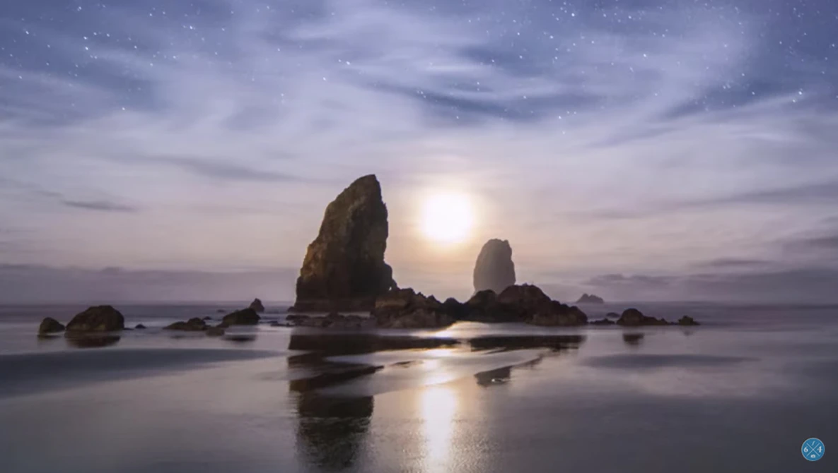 Tall rock formations silhouetted against a glowing moon rising over a calm, reflective beach at twilight.