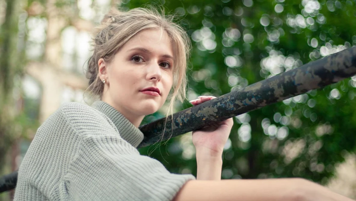 Young woman in light sweater holding weathered branch over shoulder, looking toward camera with serious expression.