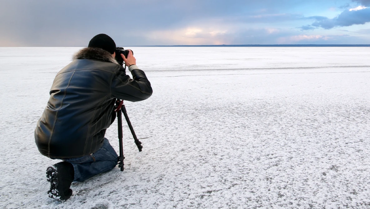 Photographer crouching on a frozen lake, composing a shot with a camera mounted on a tripod.