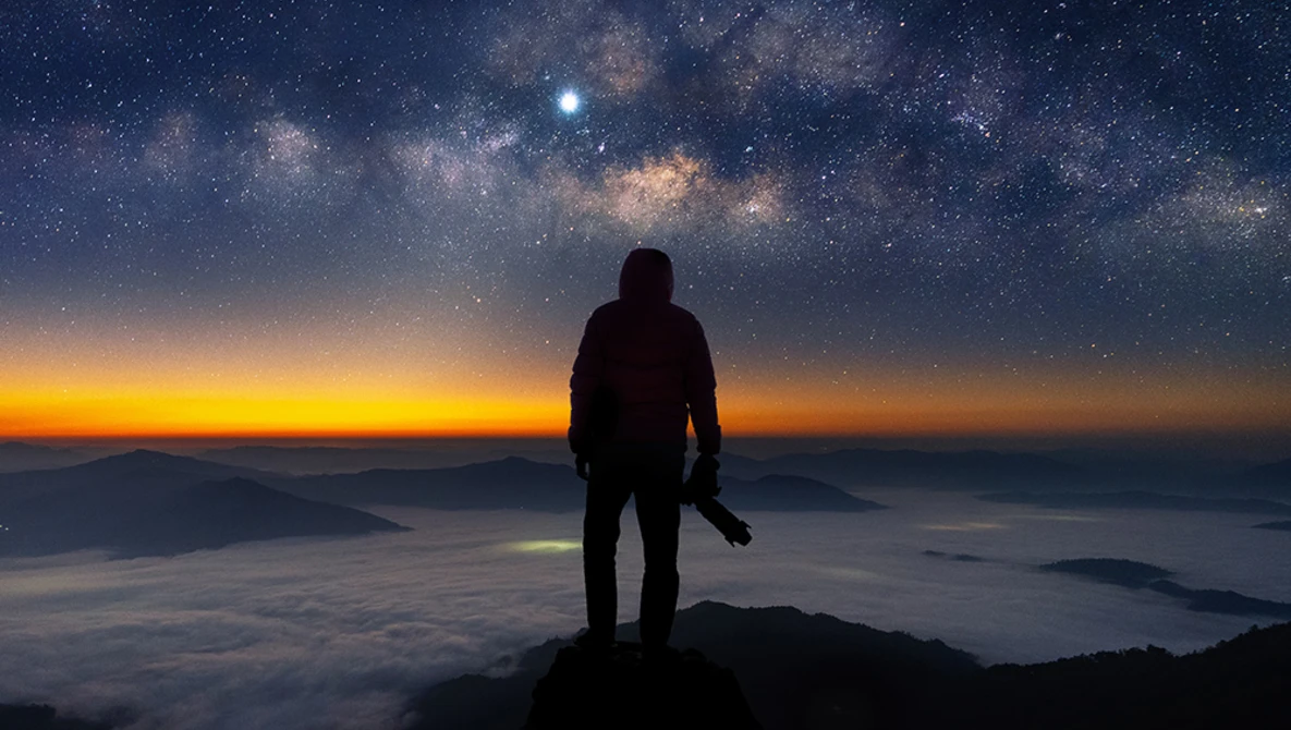 Silhouetted hiker standing on a mountain peak at sunrise, gazing toward a starry sky and Milky Way galaxy.