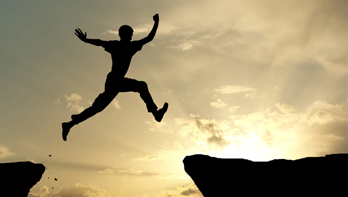 Silhouetted figure leaping between two rocky cliff edges against a golden sunset sky.