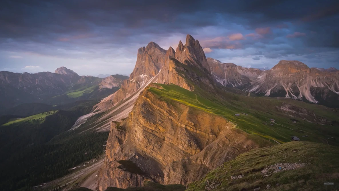 Dramatic alpine peak with steep rock faces and green meadows in a mountain valley below at golden hour.