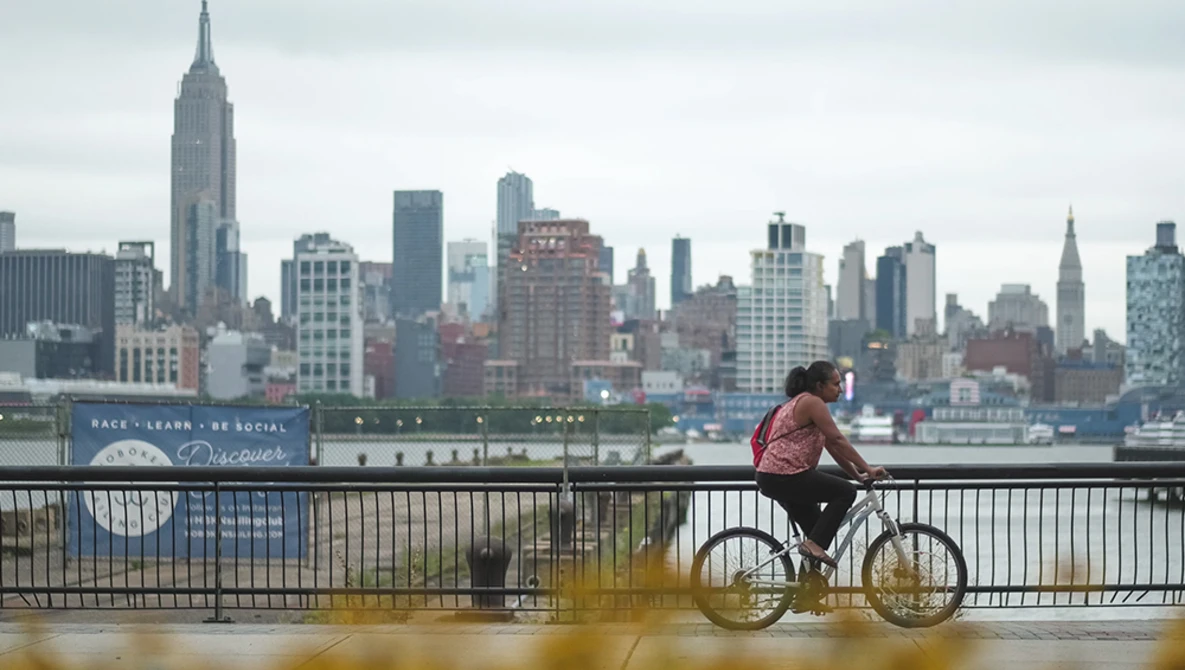 Cyclist riding a fixed-gear bike on a waterfront promenade with Manhattan skyline in background.