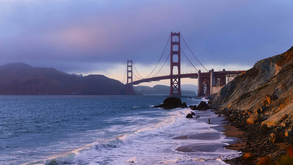 Golden Gate Bridge at dusk with rocky beach and crashing waves in foreground.