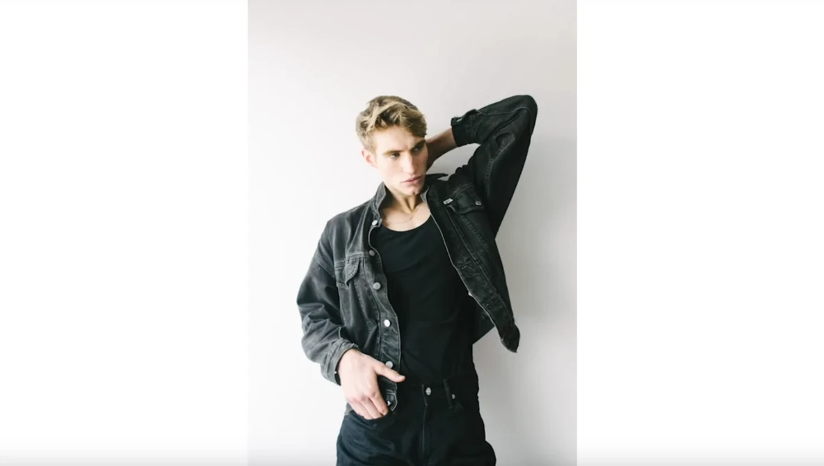 Male model in black denim jacket and shirt against white wall, hand raised to head.