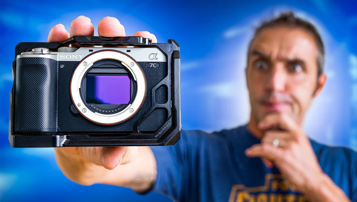 Photographer holding a mirrorless camera with visible sensor, shown against a blue sky background.