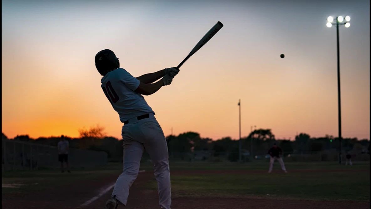 Silhouetted baseball batter mid-swing at dusk with field lights illuminated in background.