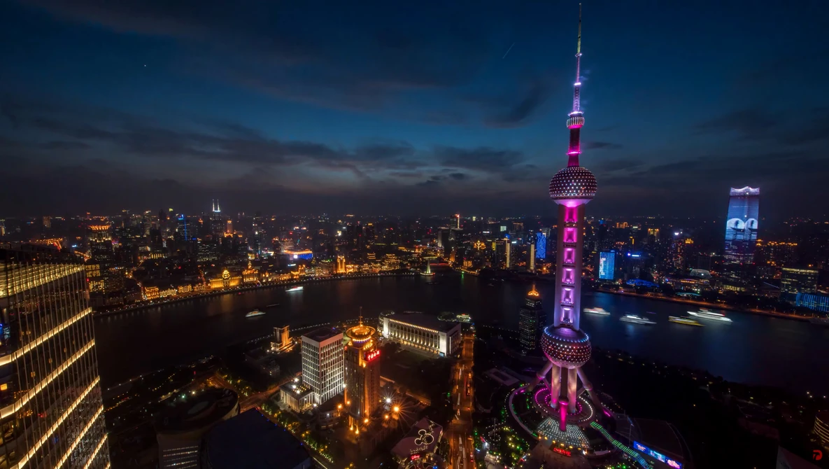 Illuminated Oriental Pearl Tower rises above Shanghai's skyline at dusk, surrounded by glowing city lights and a river.