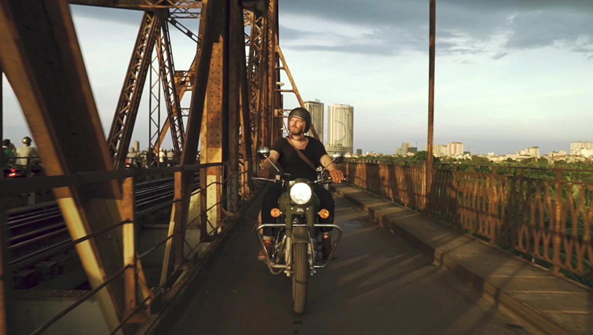 Motorcyclist riding across an industrial steel bridge toward a city skyline at golden hour.