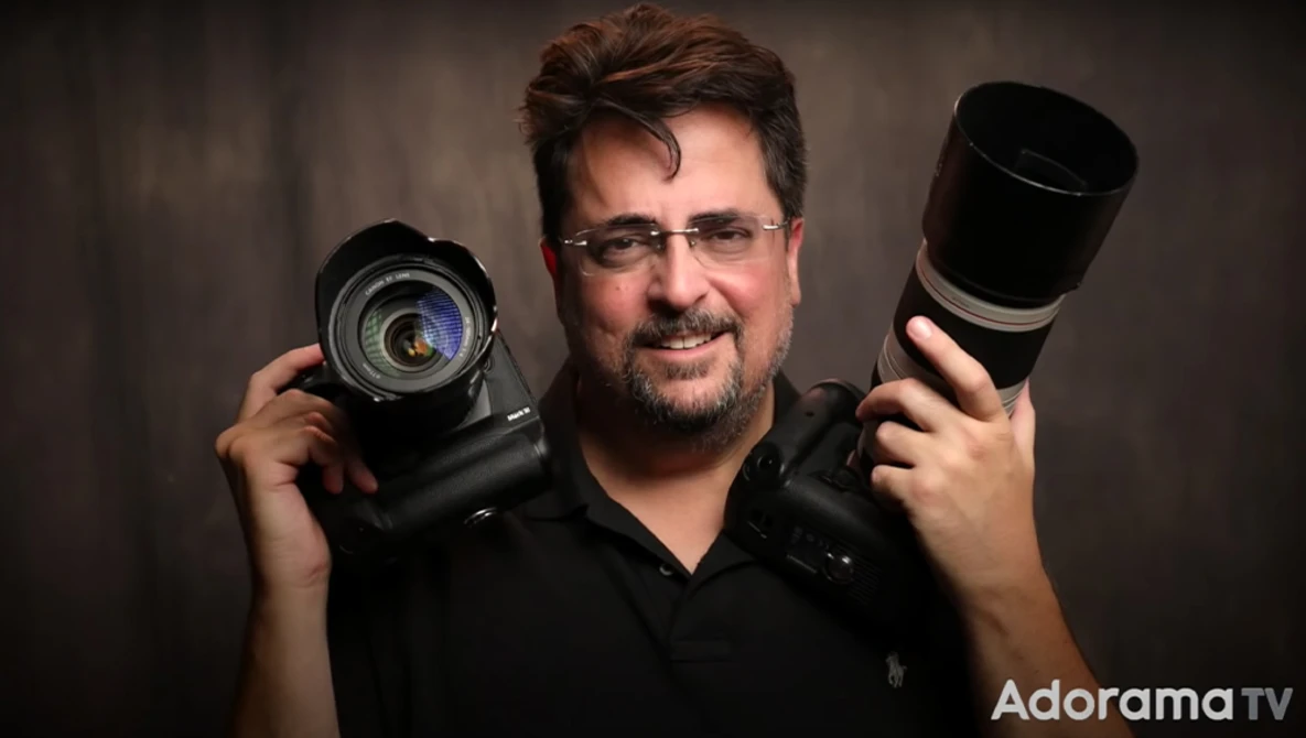 Male photographer holding a DSLR camera in one hand and a telephoto lens in the other, wearing glasses against a dark studio background.