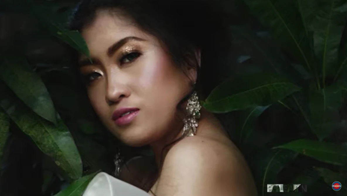 Woman with dramatic makeup and dangling earrings posed among large green leaves with side lighting.
