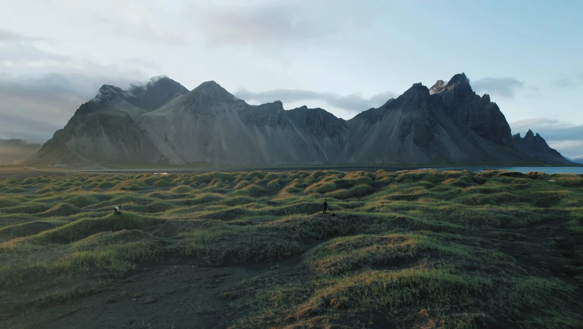 Dramatic volcanic mountain range with jagged peaks rising above a moss-covered lava field under overcast sky.