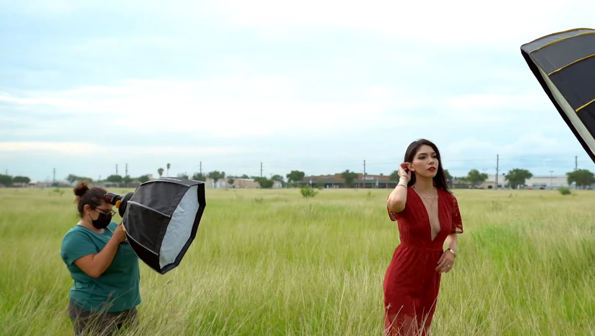 Photographer holding softbox reflector while model in red dress poses in open field.