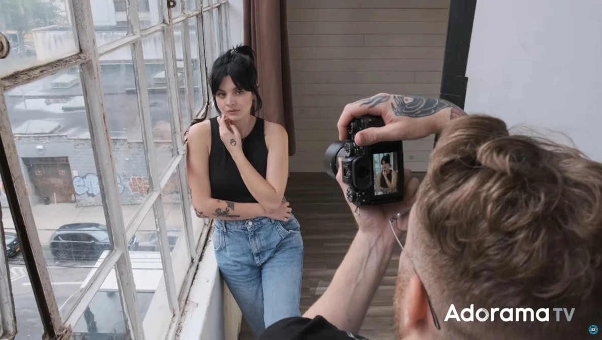 Photographer holding a camera on a tripod while shooting a portrait of a woman posing by a window.