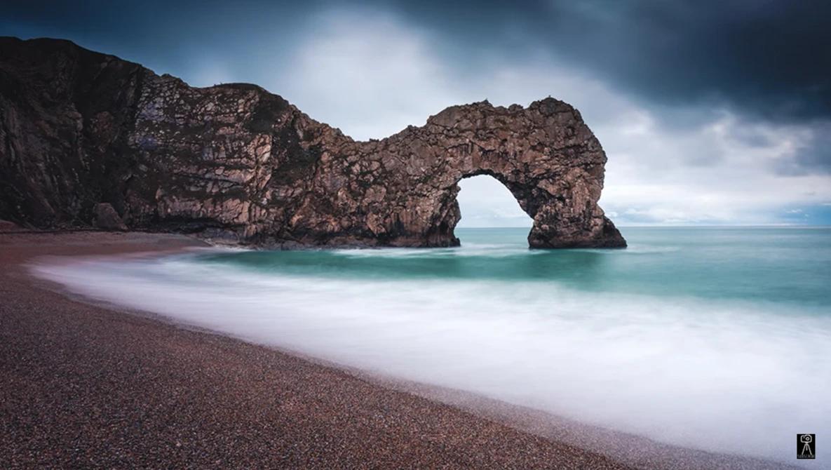 Rock arch formation in turquoise ocean waters with dark pebble beach and moody sky.