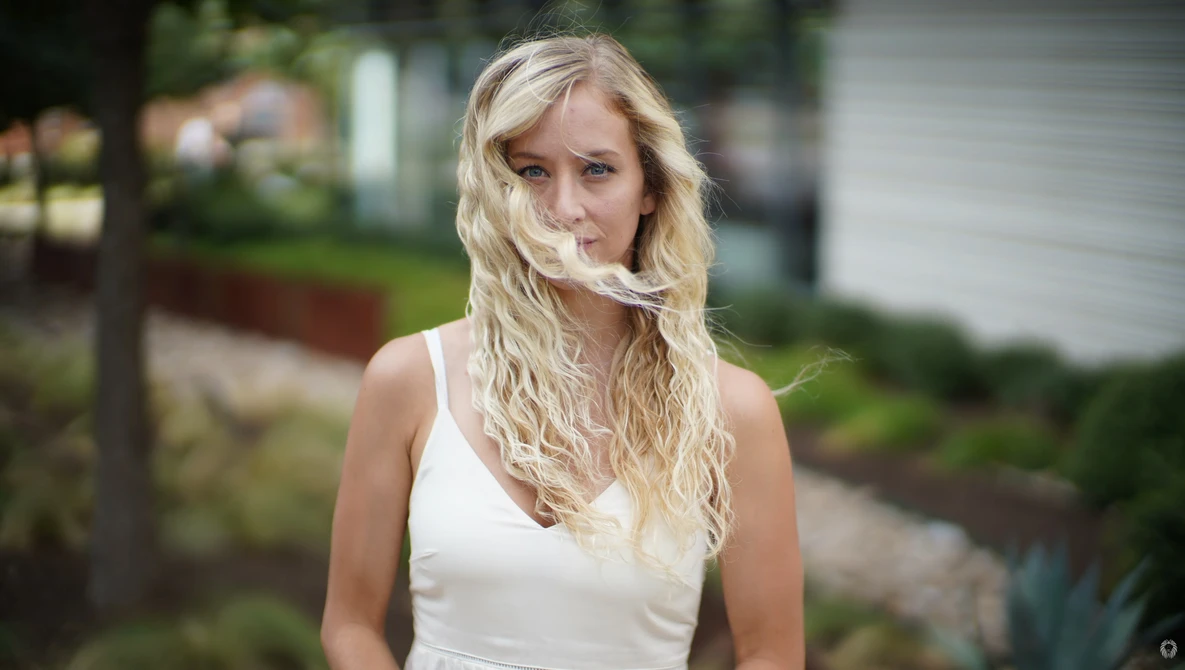 Woman with long blonde wavy hair wearing a white sleeveless top, photographed outdoors with soft natural light.
