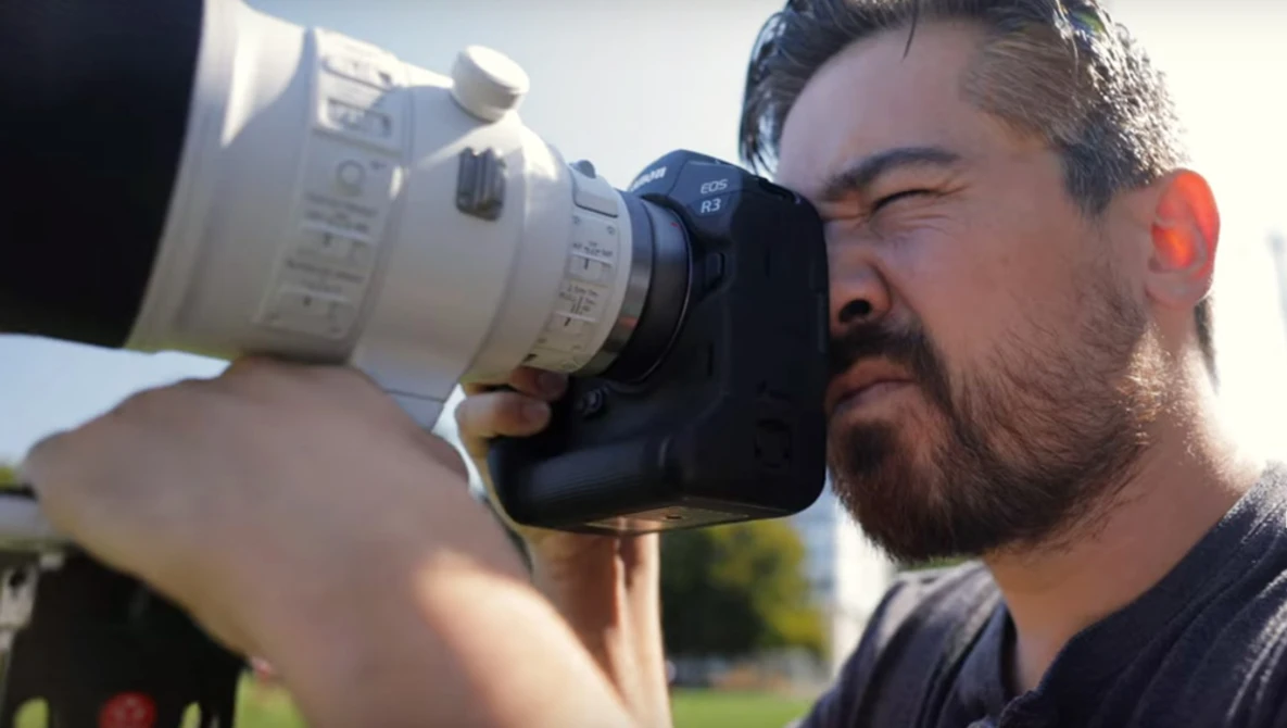 Photographer holding a telephoto lens up to his eye, framing a shot outdoors in daylight.