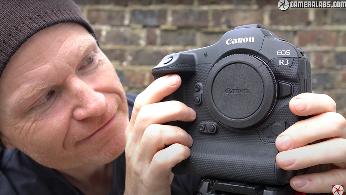 Person holding Canon EOS R3 camera body toward camera with brick wall background.