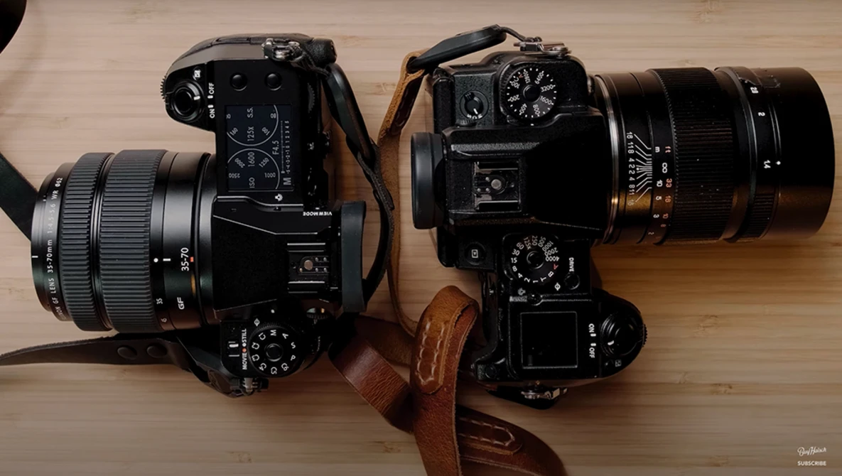 Two black film SLR cameras with attached lenses on a wooden surface, connected by a brown leather strap.