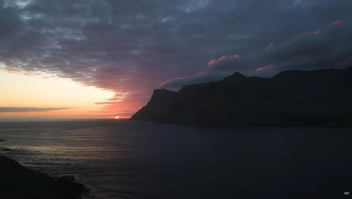 Sunset over ocean with silhouetted coastal cliffs and dramatic cloud formations.