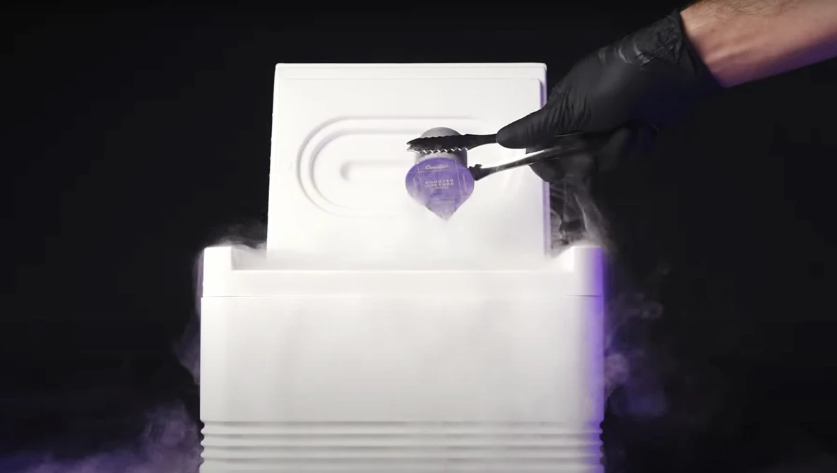 Hand in black glove holding tweezers over a purple spherical object suspended above a white textured surface with concentric circular patterns.