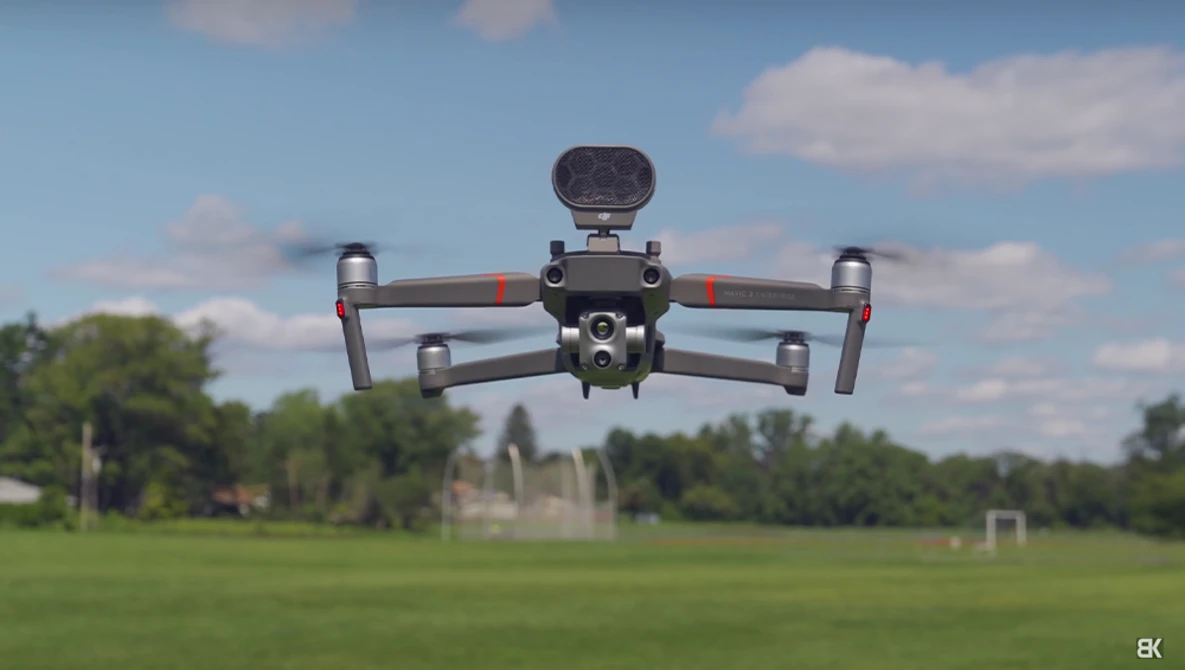 DJI Mavic 3 drone hovering above a green field with trees and buildings in the background.