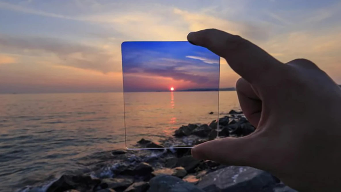 Hand holding a transparent glass panel framing a sunset over the ocean with rocky shore in foreground.