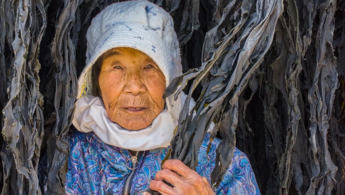 Elderly woman wearing a white head covering and blue patterned garment stands before a weathered backdrop of dried palm fronds or similar fibrous material.