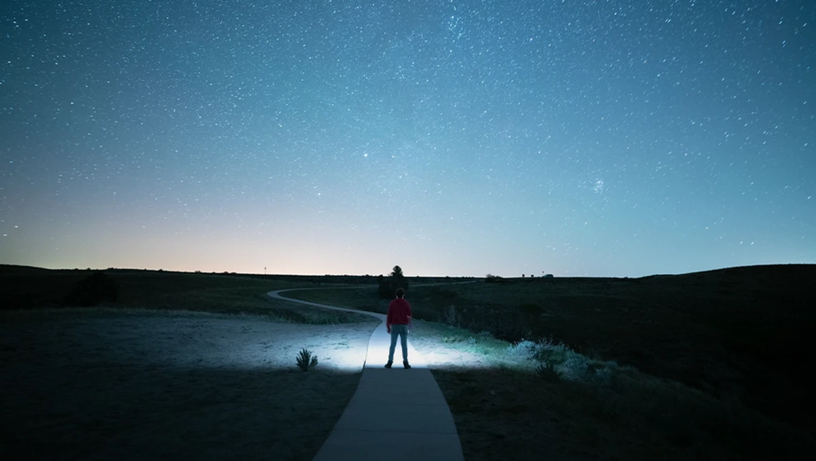 Silhouetted figure standing on a boardwalk, illuminated by headlamp light, facing a starry night sky over a dark landscape.