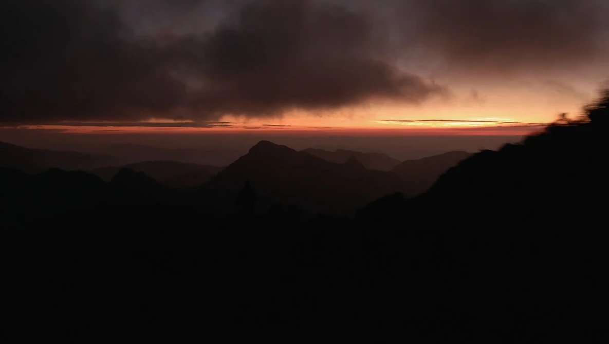 Silhouetted mountain ridges against an orange and gold sunset sky with dramatic clouds.
