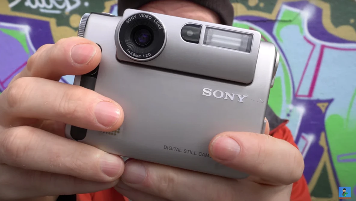 Person holding a silver Sony digital still camera with a compact design and retractable lens.